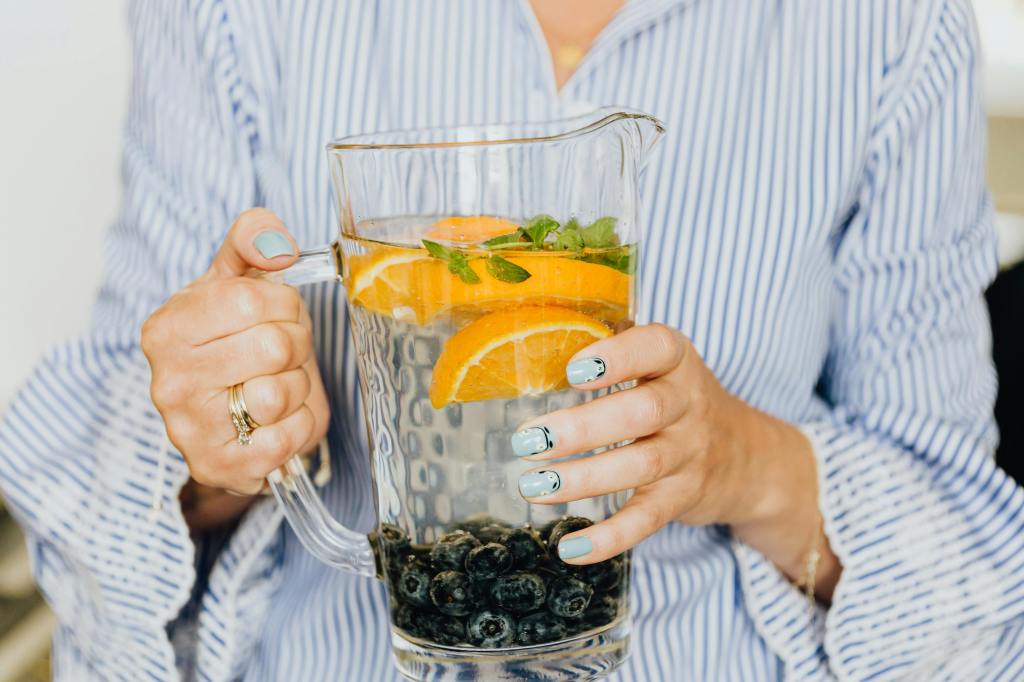 A woman holding a pitcher of water with orange slices, blueberries and a sprig of mint. Noticeable absent is no sugar